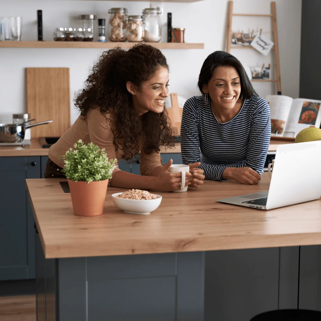 Dos mujeres en una cocina revisando informacion en un ordenador, planificando el cambio de denominacion social de su negocio.