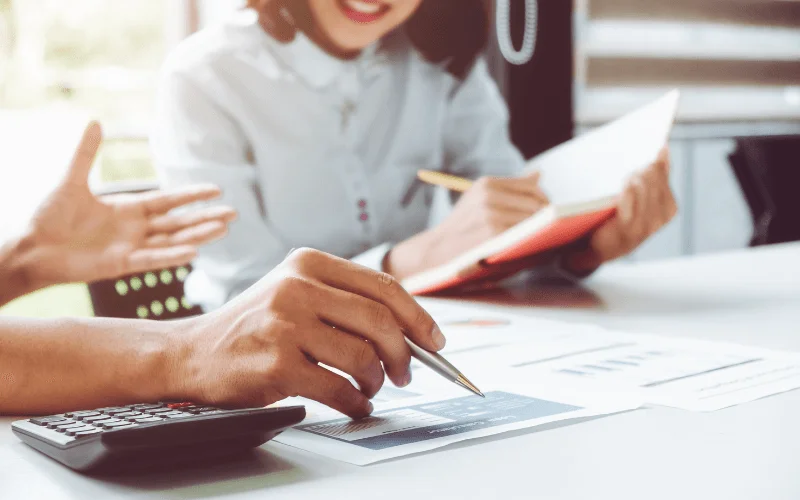 Manos revisando graficos financieros y calculadora, mientras dos personas analizan estrategias para ampliaciones y reducciones de capital online.
